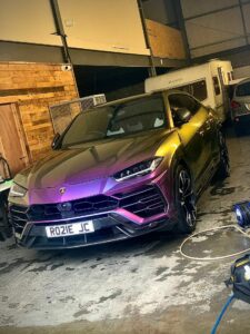 A metallic chameleon-colored Lamborghini Urus is parked in a dimly lit garage. The SUV has a custom license plate "RO21E JC" and is surrounded by various equipment and other vehicles, including a camper van and a car partially visible in the background.