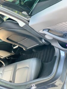 Interior of a car, viewed from the open passenger-side rear door. The image shows the back seats, black carpeting, and part of the front passenger seat. The car door interior features a speaker and a small storage compartment. The upholstery is grey and black.