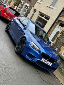 A blue BMW parked on a residential street with terraced houses. Another red car is parked in front. The street appears wet, suggesting recent rain. House numbers and license plates are visible.