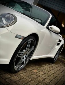 A close-up shot of a white sports car parked on a brick driveway. The image focuses on the car's left front side, showcasing the sleek body design, detailed rims, and low-profile tires, with part of the background including a dark open garage door.