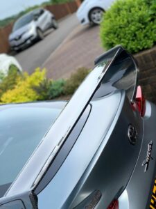 Close-up view of the rear end of a grey car featuring a large carbon fiber spoiler. The car is parked on a paved driveway, with another car and well-maintained bushes in the background.