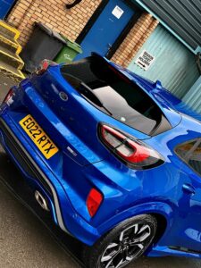 A blue Ford Puma parked in front of a brick building with a blue door. A green garbage bin is next to the door, which has a sign reading "HELSTON GARAGE HOLBORN" above it. The vehicle’s registration plate reads "EG22 RTX.