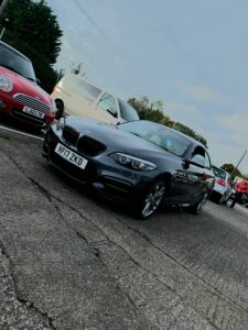 A sleek, dark gray BMW with the license plate "RF17 ZKD" is parked in a lot with other cars, including a red Mini Cooper and a white SUV. The sky is clear with a few clouds, and trees are visible in the background.