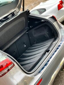 Open trunk of a modern silver car, showing a spacious and empty storage area with a black interior lining. The car is parked on a street with part of a white car visible in the background. The trunk lid is raised, revealing the clean and organized space.