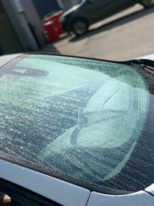 A close-up of a car windshield covered in raindrops during daylight. Sunlight glistens off the water droplets, creating a shimmering effect. The car's hood and part of the interior are visible through the glass. A blurred background includes another car and a trash bin.