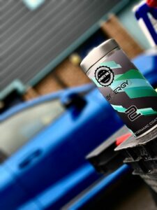 A silver and teal canister labeled "Synergy" is perched on a black surface in the foreground, with a blurred blue car and brick building in the background. The canister appears to be a car care product, possibly for ceramic coating.