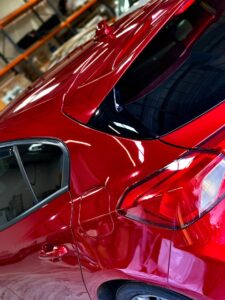 A close-up view of the rear section of a shiny red car, highlighting the rear window, taillight, and fuel cap. The indoor setting shows some shelves and boxes in the background, slightly out of focus.