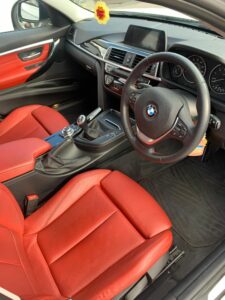 Interior view of a luxury car featuring red leather seats, a BMW emblem on the steering wheel, a modern dashboard with a digital display, and silver trim accents. The center console includes a gear shift, control knobs, and a flower-shaped air freshener hanging.