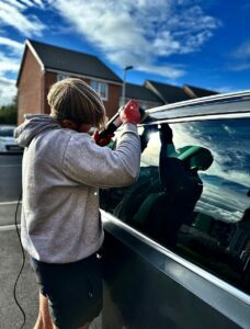 A person with short hair, wearing a hoodie and shorts, fixes a device on the side window of a vehicle. They are outdoors in a parking lot near residential houses. The sky is blue with scattered clouds, and the sun is shining brightly.