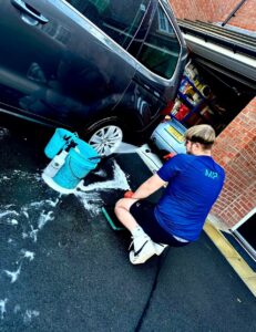 A man kneels on the driveway near a bucket filled with soapy water, washing the wheel of a dark-colored car parked outside a garage. The garage door is open, exposing shelves filled with various items. The man wears a blue T-shirt and black shorts.