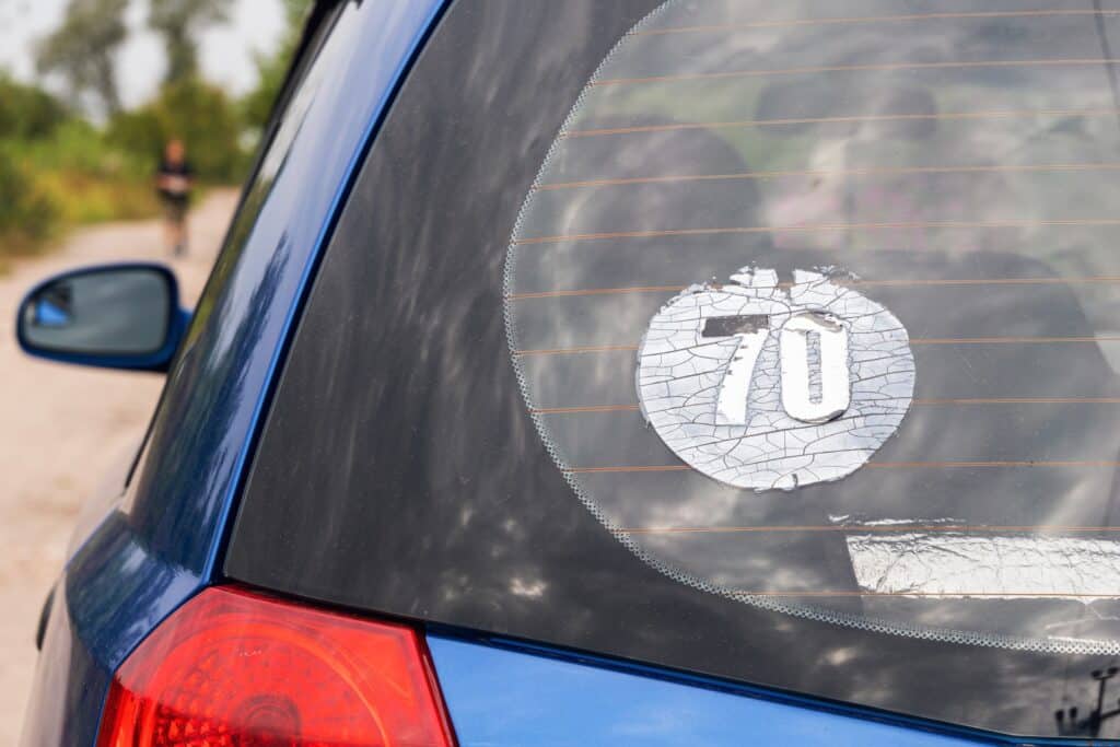 A blue car with a cracked circle sticker on the rear window showing the number 70. The background is blurred, with greenery and a person walking in the distance.