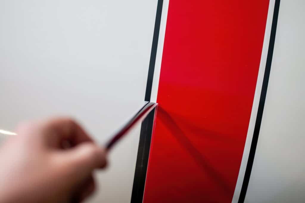 A hand holding a tool is about to remove or apply a red and black striped decal from a white surface.