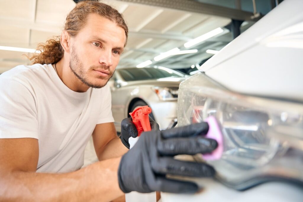 A man wearing a white shirt and black gloves is polishing a car headlight with a pink cloth. A spray bottle is held in his other hand. The setting appears to be a well-lit garage or workshop.