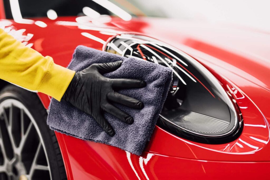 A person wearing a yellow sleeve and black glove uses a gray microfiber cloth to polish the headlight of a shiny red sports car. The image focuses on the detailing work on the vehicle's front.