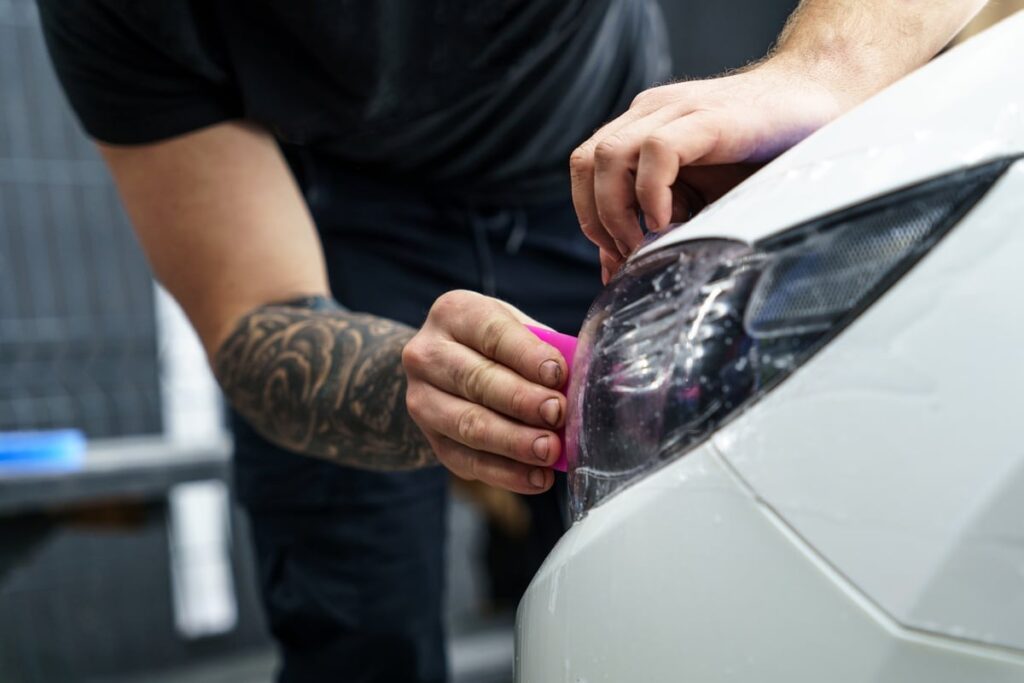 A person with a tattooed forearm is applying a protective film to a car's headlight using a pink tool. The car is white, and the focus is on the careful detailing of the headlight area.